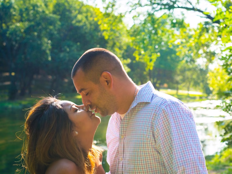 A couple about to kiss by the water, sunlit trees behind them